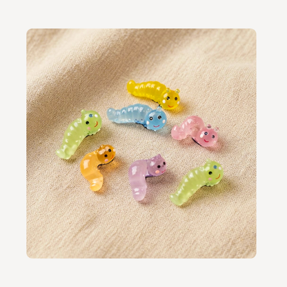Seven small translucent resin caterpillar clog charms in pastel colors, including yellow, blue, pink, orange, purple, and light green, arranged on a textured beige fabric background.