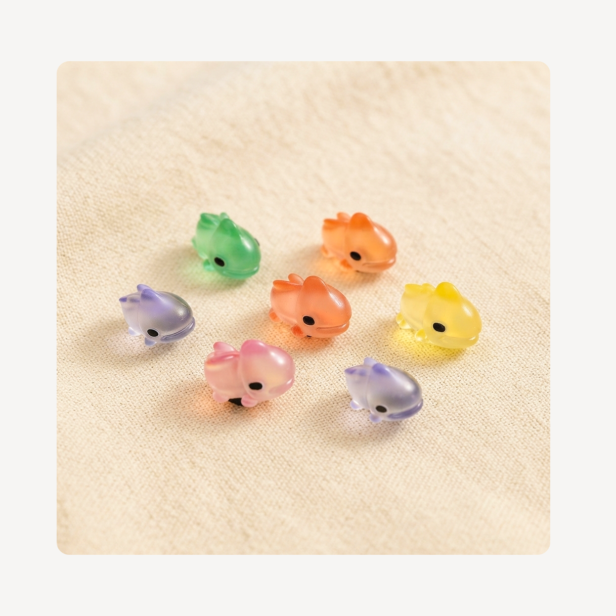 Seven small translucent chameleon-shaped clog charms in pastel colors, including green, orange, yellow, pink, and lavender, arranged on a textured beige fabric background.