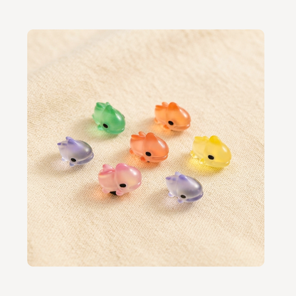 Seven small translucent chameleon-shaped clog charms in pastel colors, including green, orange, yellow, pink, and lavender, arranged on a textured beige fabric background.