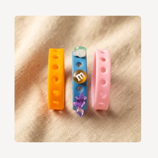 Three silicone clog charm wristbands with hole details are displayed on a textured beige fabric background. The wristbands are orange, light blue, and pink, with decorative charms including a silver glitter cloud, a gold M&M's coin, and a purple flower.