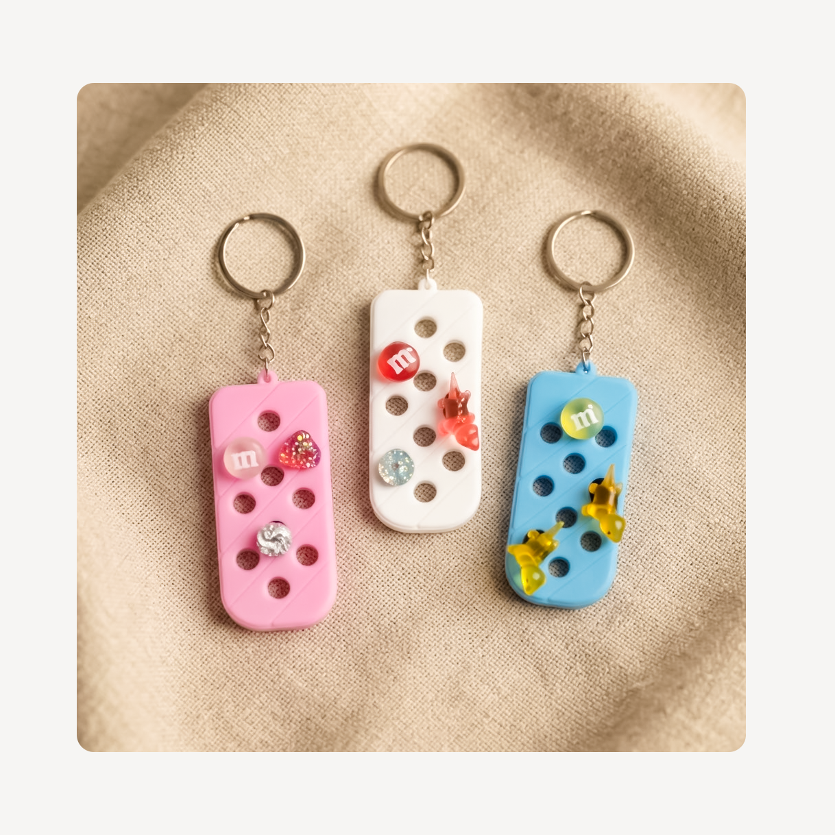 Three rectangular clog charm keychains with metal key rings are arranged on a beige fabric background. Each keychain has a small colored charm with a circular cutout and a tiny red M-shaped accent.