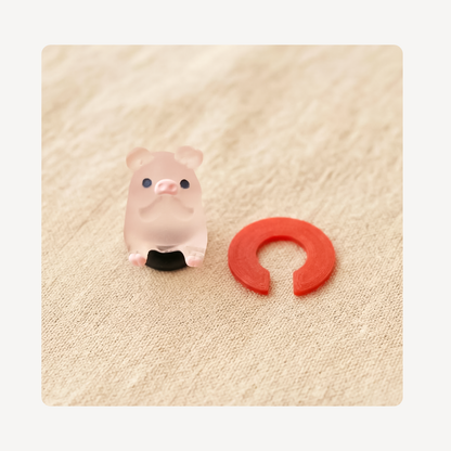 A small translucent pale pink pig clog charm with black feet and black eyes sits beside a red U-shaped ring on a beige textured fabric background.