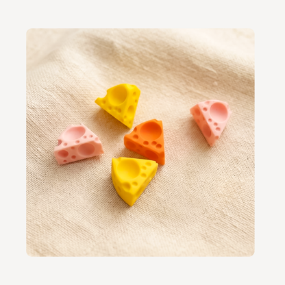 Five mini cheese-shaped clog charms in pastel pink, orange, and yellow, arranged on a textured beige linen fabric background.