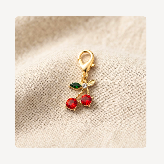 A gold shoe charm shaped like two cherries, featuring red faceted gemstones, green leaves, and a small clear rhinestone, resting on a textured beige linen fabric background.