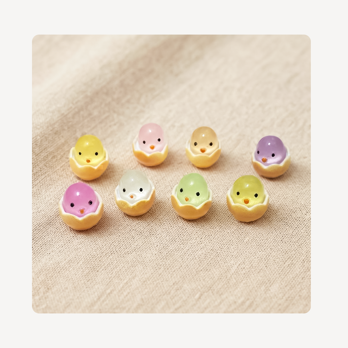 Seven small pastel chick-shaped clog charms arranged on a beige textured fabric background. Each chick is cracked open at the top, showing a soft pastel body color in yellow, pink, peach, purple, white, green, and yellow.
