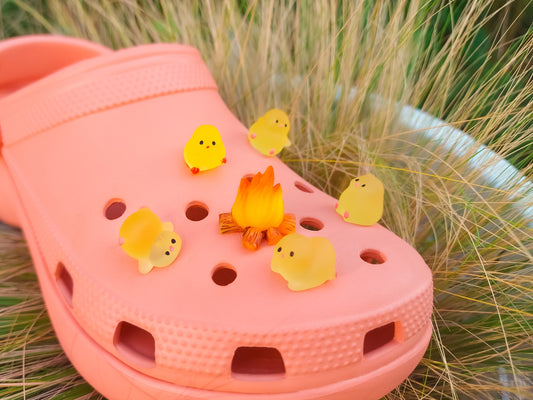 A pink clog-style shoe decorated with small yellow chick figurines and a tiny orange campfire, placed on green grass.