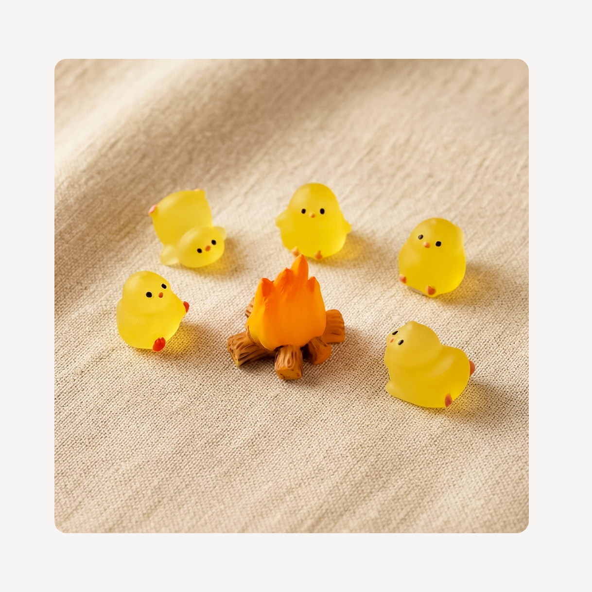 Five small translucent yellow chick  clog charms with simple black eyes and red feet are arranged around a tiny orange campfire on a beige textured fabric background.