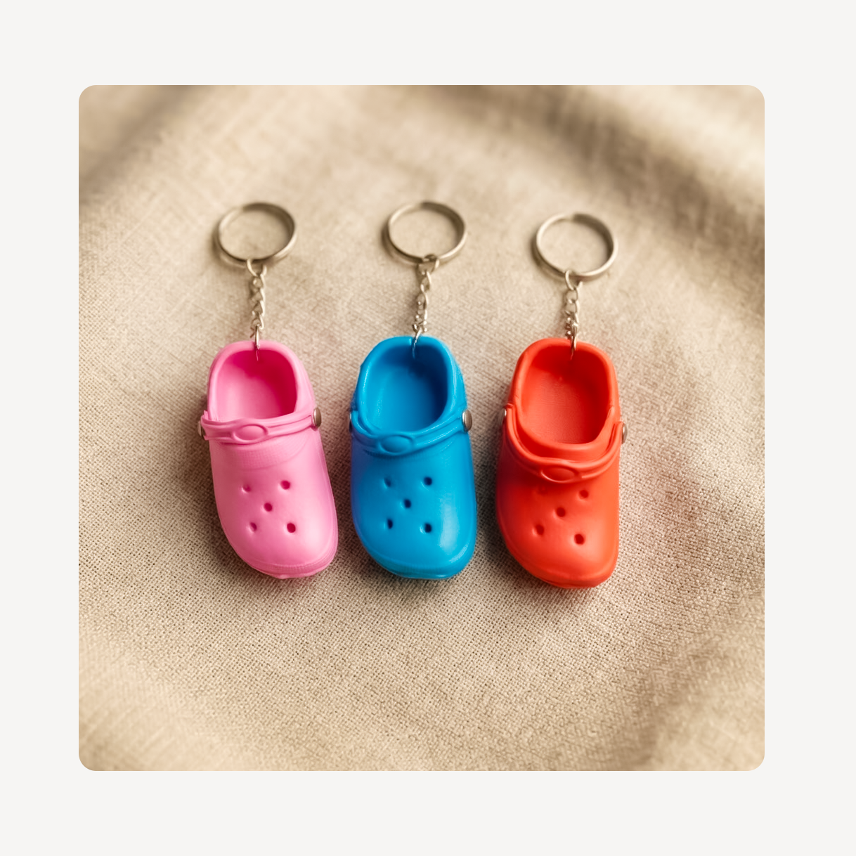 Three miniature clog-shaped keychains in pink, blue, and orange, arranged side by side on a textured beige fabric background.