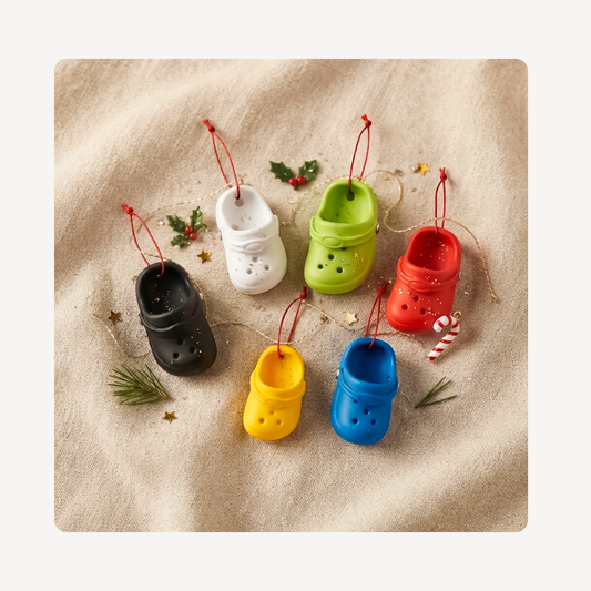 Five miniature clog-shaped Christmas ornaments in black, white, green, yellow, and blue, each with a red hanging loop, arranged on a beige fabric background with small holiday props.