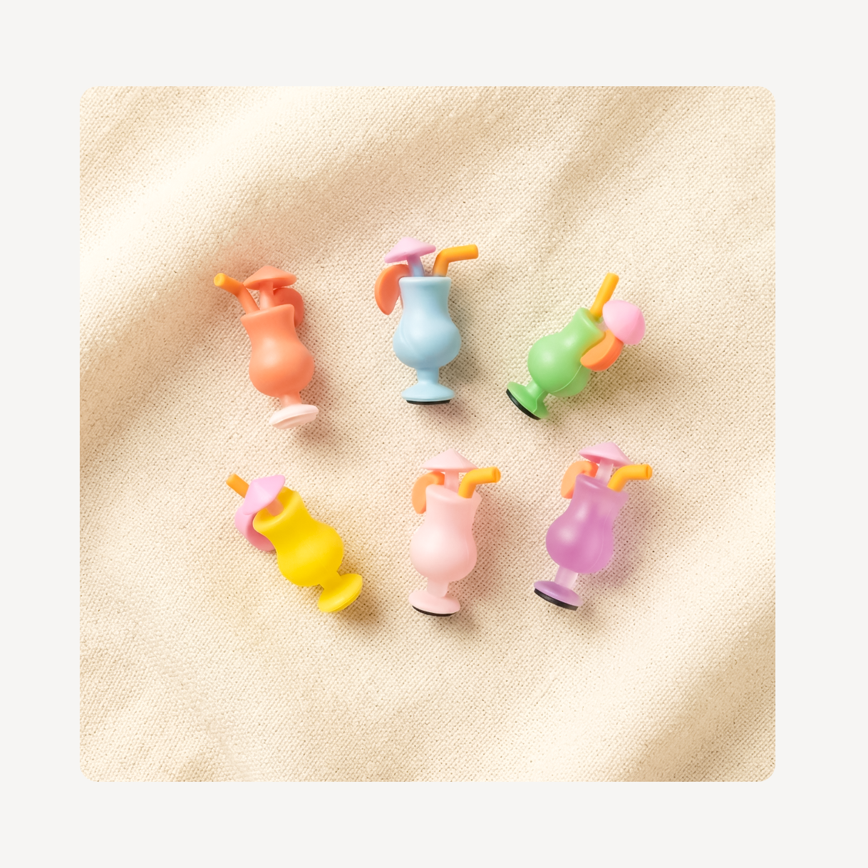 Six small pastel cocktail-shaped clog charms arranged on a soft beige fabric background. The set includes orange, light blue, green, yellow, pink, and purple pieces with orange straws and small black circular accents.