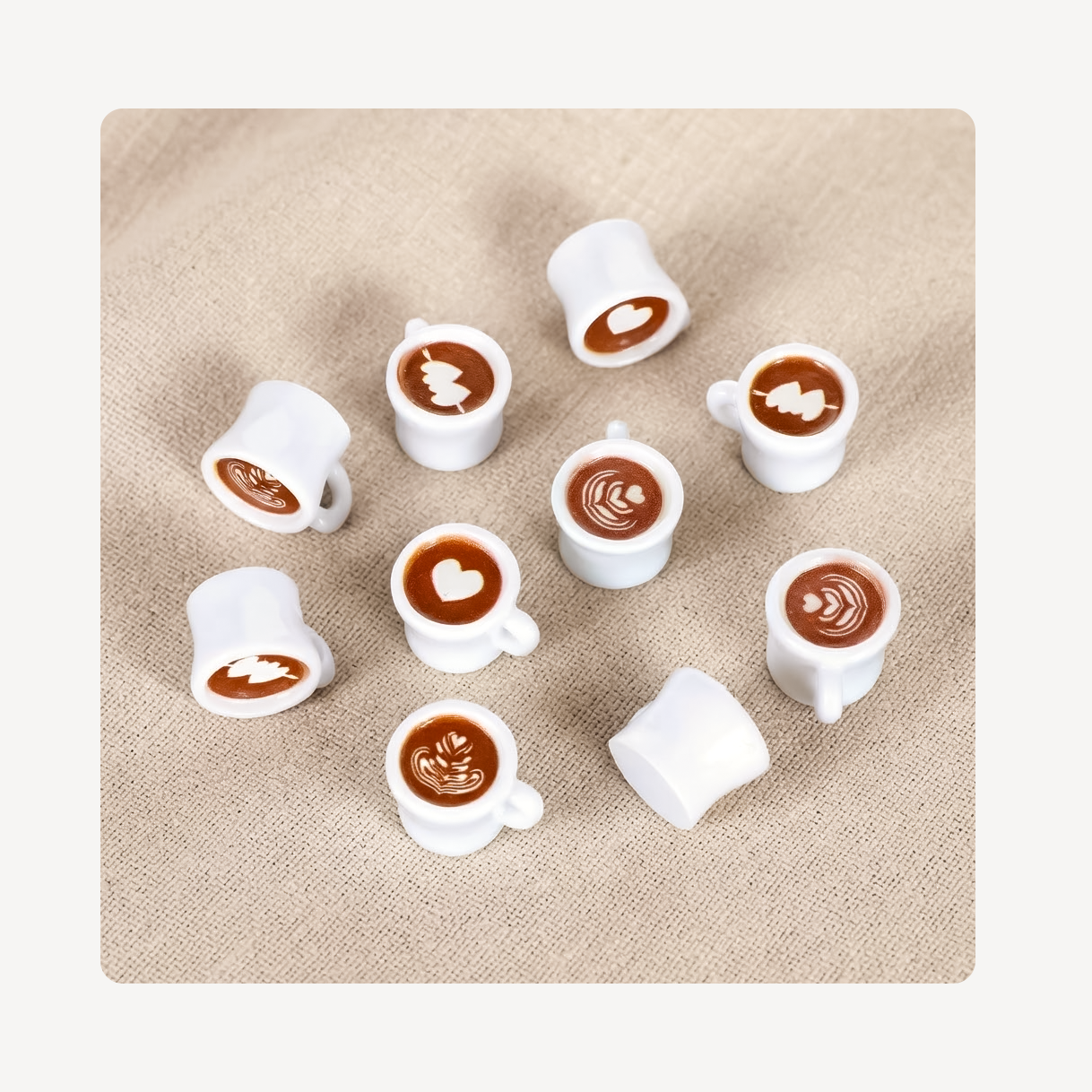 Nine miniature white ceramic coffee cup clog charms with various latte art designs, arranged on a textured beige fabric background.