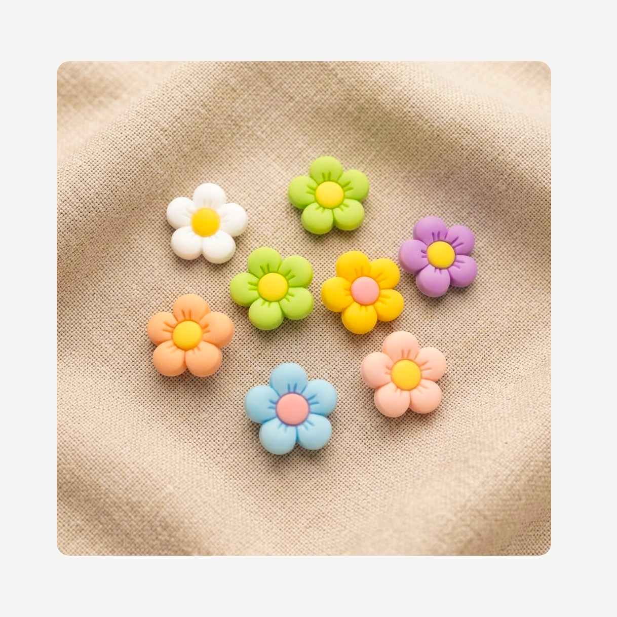 Eight small flower-shaped clog charms in pastel colors, including white, green, yellow, purple, orange, blue, and pink, arranged on a beige textured fabric background.