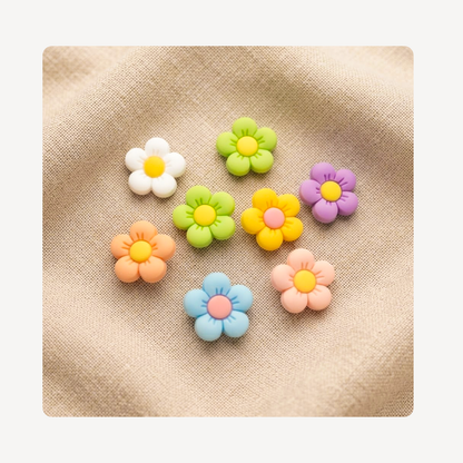 Eight small flower-shaped clog charms in pastel colors, including white, green, yellow, purple, orange, blue, and pink, arranged on a beige textured fabric background.