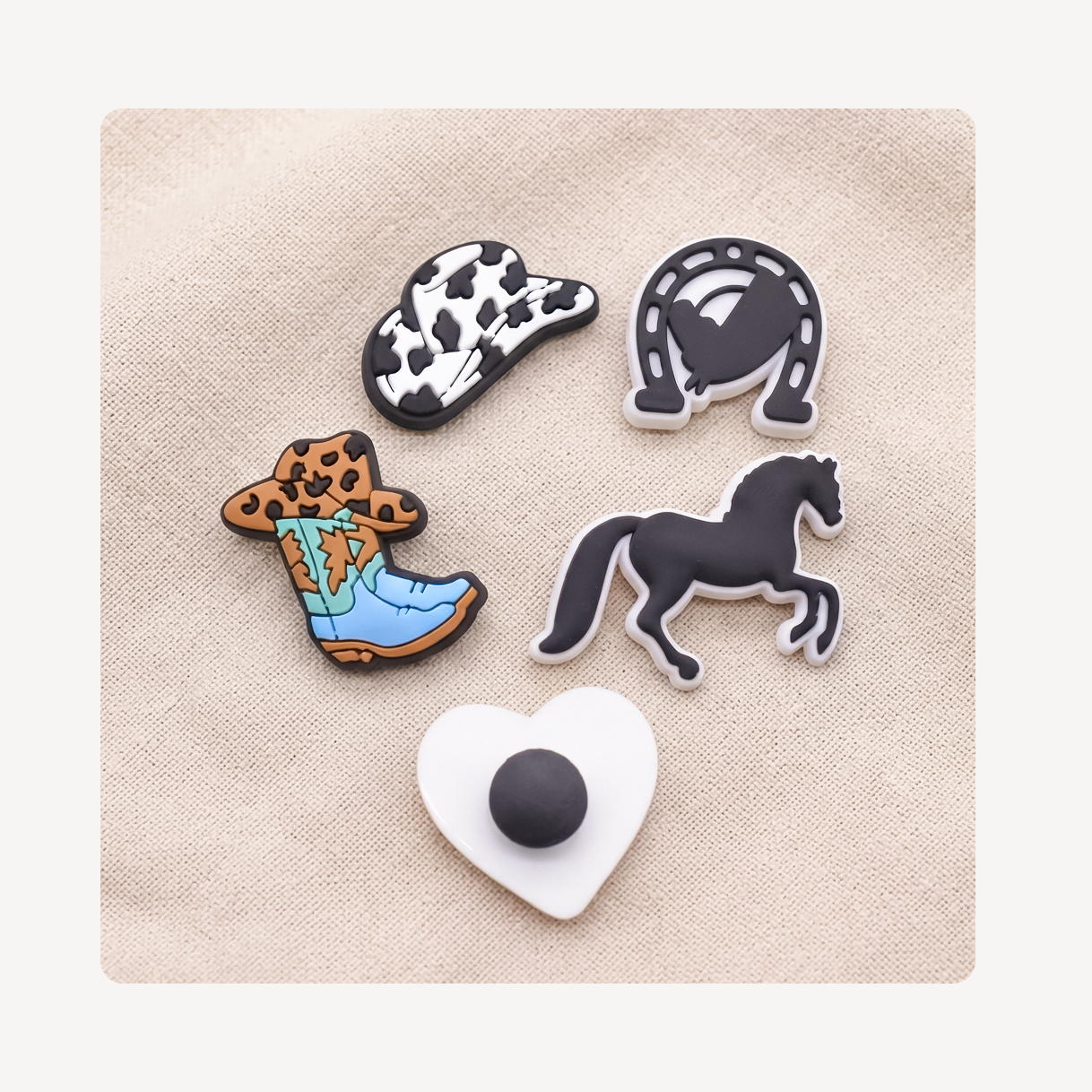 Five cow-themed decorative clog charms arranged on a beige fabric background: a black-and-white cow-print hat, a horseshoe, a cowboy boot with leopard print and blue detailing, a black horse silhouette, and a white heart-shaped piece with a black center.