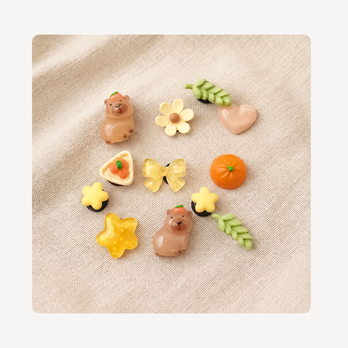 A set of nine cute, small decorative clog charms arranged on a textured beige linen background. The charms include two brown bears, a yellow flower, a pink heart, green leaves, an orange pumpkin, a yellow butterfly, a yellow flower with a black center, a yellow star, a yellow flower with a green center, and a triangular pastry with a strawberry on top.