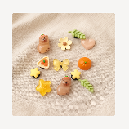 A set of nine cute, small decorative clog charms arranged on a textured beige linen background. The charms include two brown bears, a yellow flower, a pink heart, green leaves, an orange pumpkin, a yellow butterfly, a yellow flower with a black center, a yellow star, a yellow flower with a green center, and a triangular pastry with a strawberry on top.