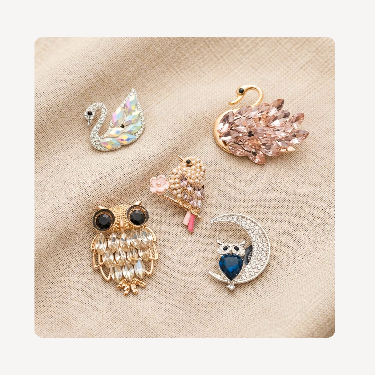 Five decorative clog charms arranged on a beige textured fabric background: a silver swan with iridescent stones, a pink feathered swan, a gold owl with black eyes and white feather details, a small gold bird with pink accents, and a crescent moon brooch with a blue heart-shaped face and white crystal border.