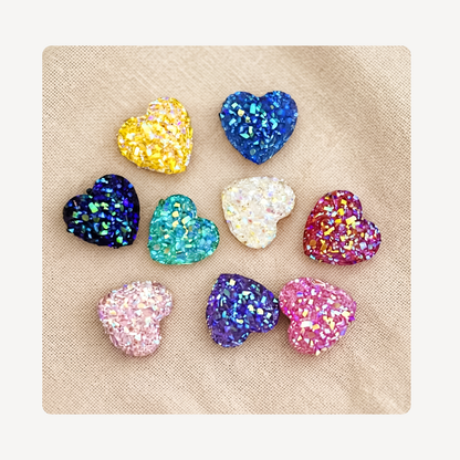 Nine glittery heart-shaped clog charms in various colors arranged on a beige textured fabric background.