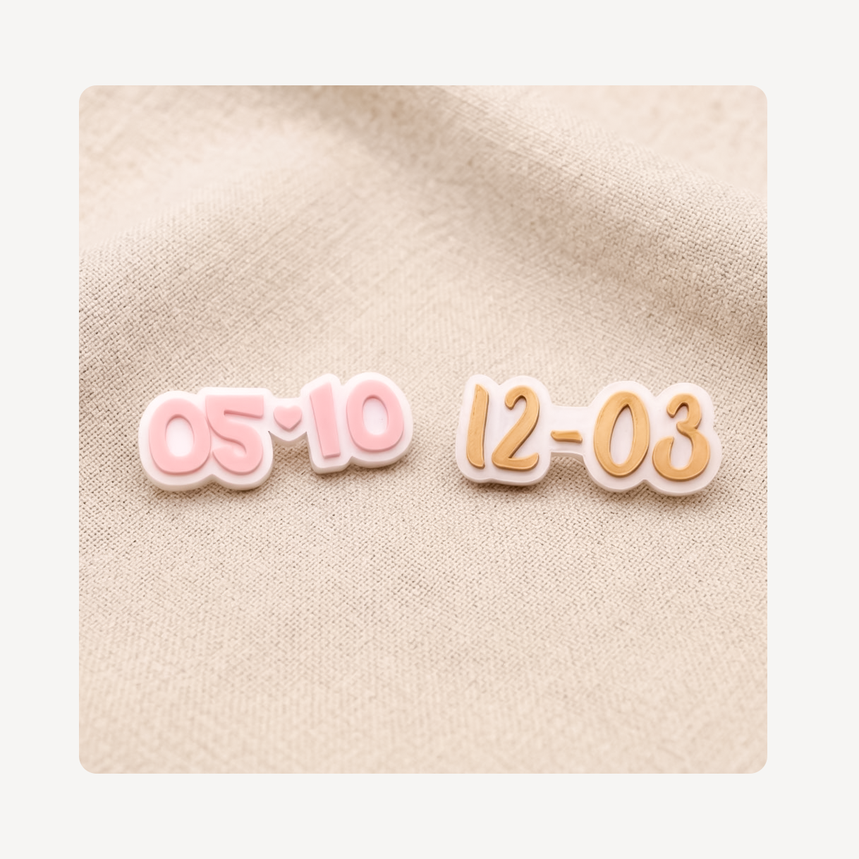 Two small date-shaped clog charms on a beige fabric background, one in pink outlined in white and the other in peach-orange outlined in white.