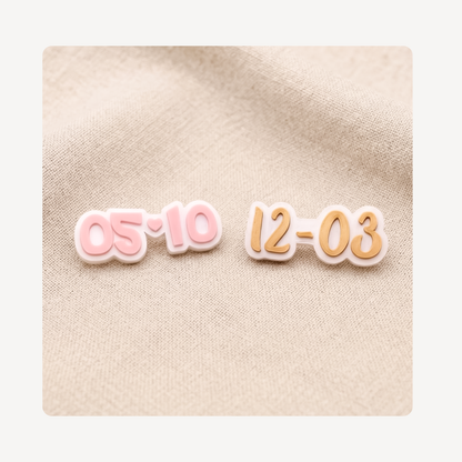 Two small date-shaped clog charms on a beige fabric background, one in pink outlined in white and the other in peach-orange outlined in white.