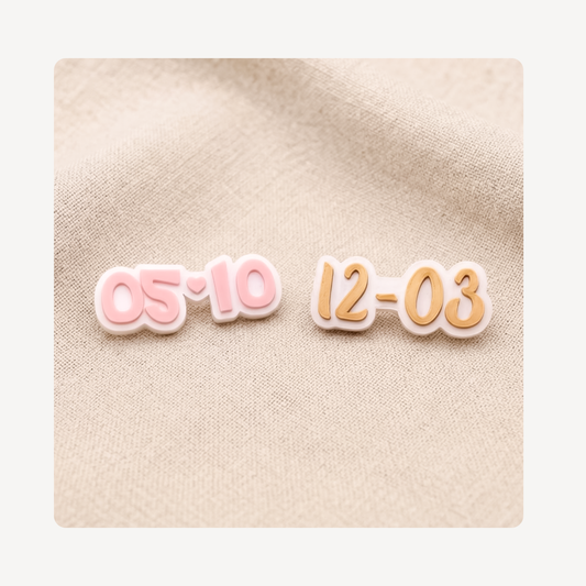 Two small date-shaped clog charms on a beige fabric background, one in pink outlined in white and the other in peach-orange outlined in white.