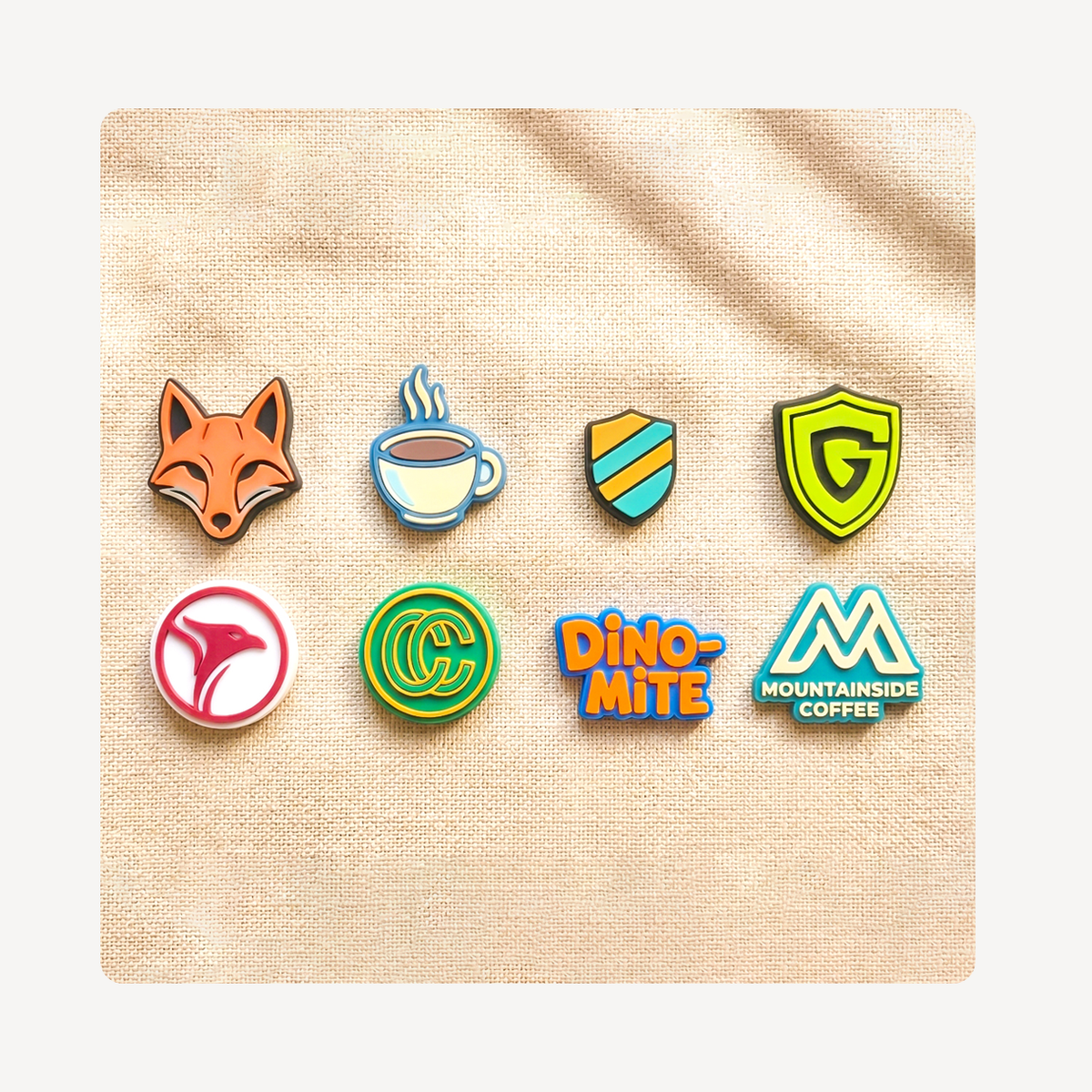 Eight colorful custom logo clog charms arranged in two rows on a textured beige fabric background. The charms feature various designs including a fox head, a coffee cup, shield emblems, a pink abstract shape, a green circular logo, orange and blue 'DINO-MITE' text, and a blue and white 'MOUNTAINSIDE COFFEE' logo.