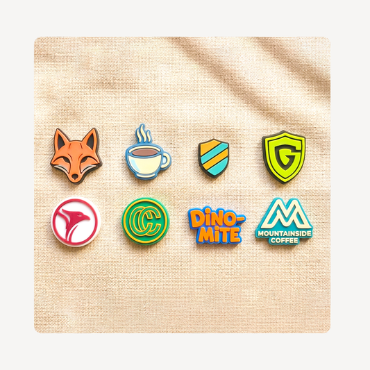 Eight colorful custom logo clog charms arranged in two rows on a textured beige fabric background. The charms feature various designs including a fox head, a coffee cup, shield emblems, a pink abstract shape, a green circular logo, orange and blue 'DINO-MITE' text, and a blue and white 'MOUNTAINSIDE COFFEE' logo.