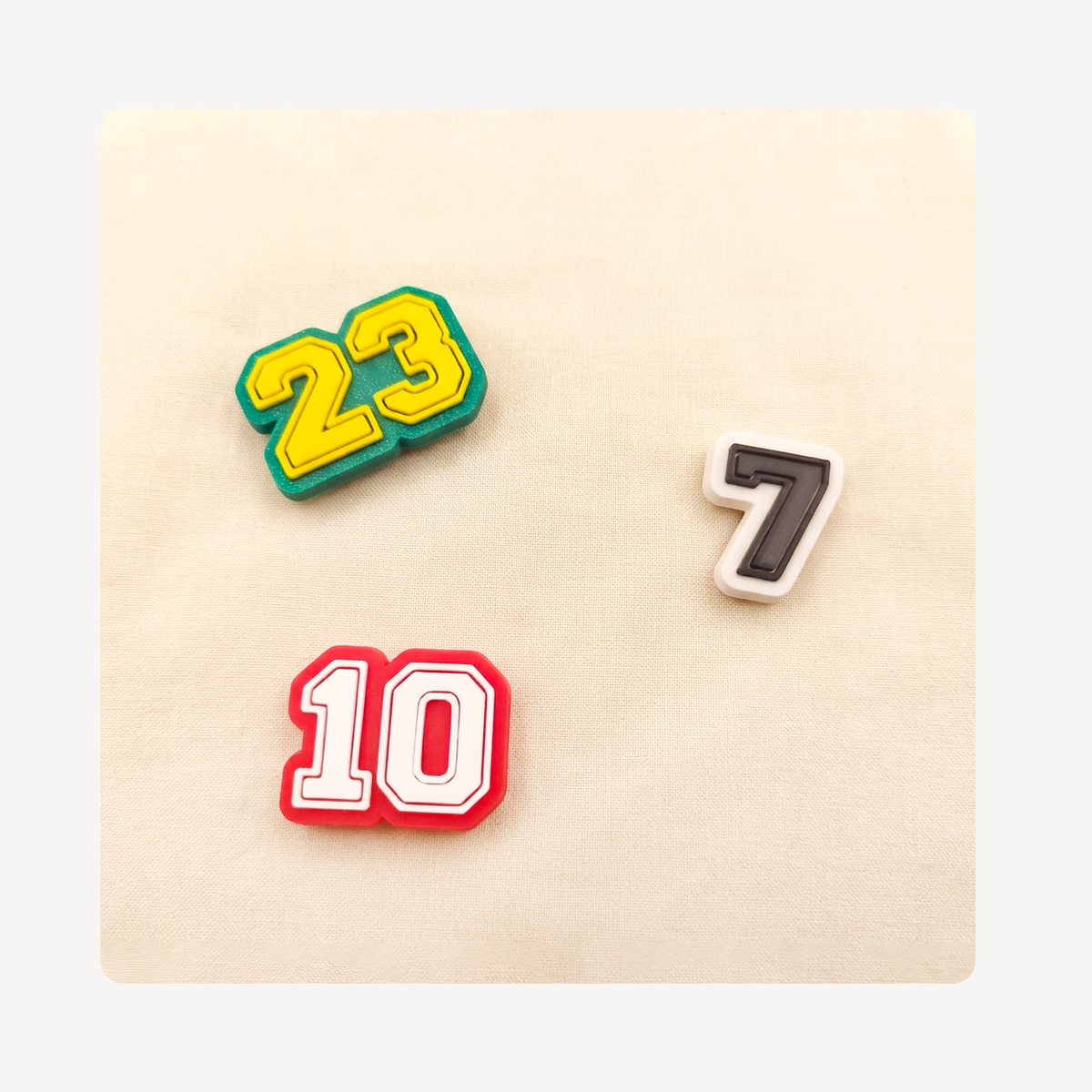 Three athletic-style number clog charms, including 23 in green and yellow, 10 in red and white, and 7 in black and white, arranged on a beige textured background.
