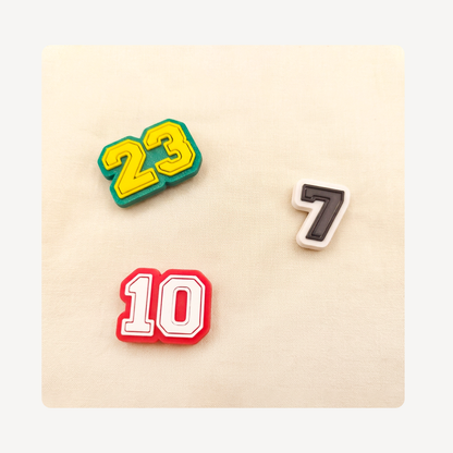 Three athletic-style number clog charms, including 23 in green and yellow, 10 in red and white, and 7 in black and white, arranged on a beige textured background.