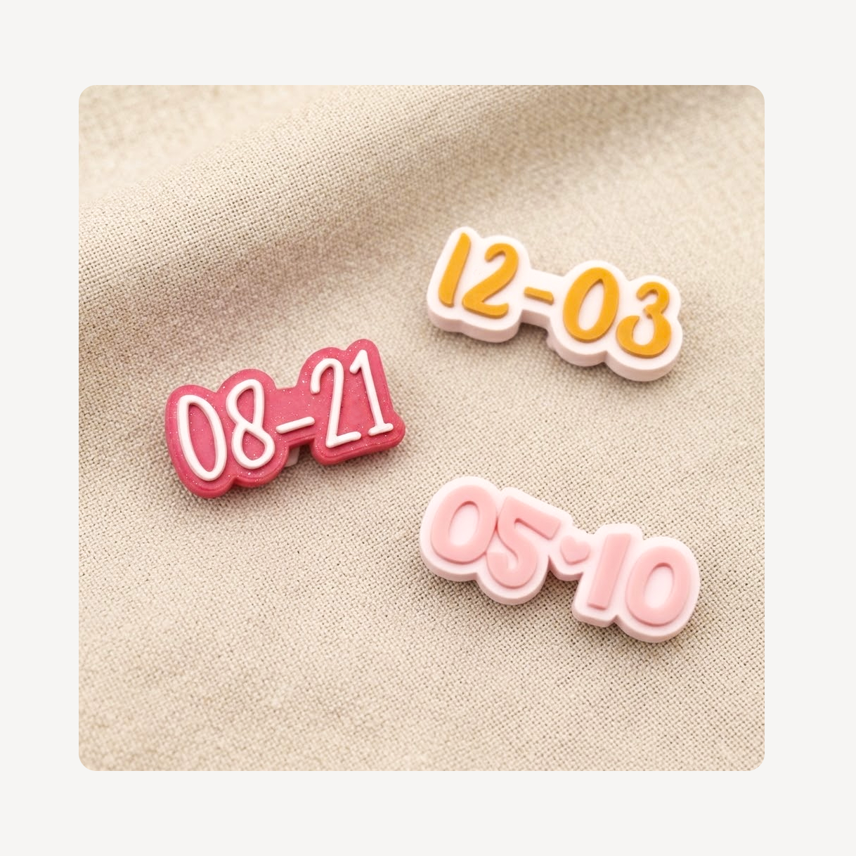 Three small date-number shoe charms reading 08-21, 12-03, and 05-10 in pink, red, and yellow tones, arranged on a beige fabric background.