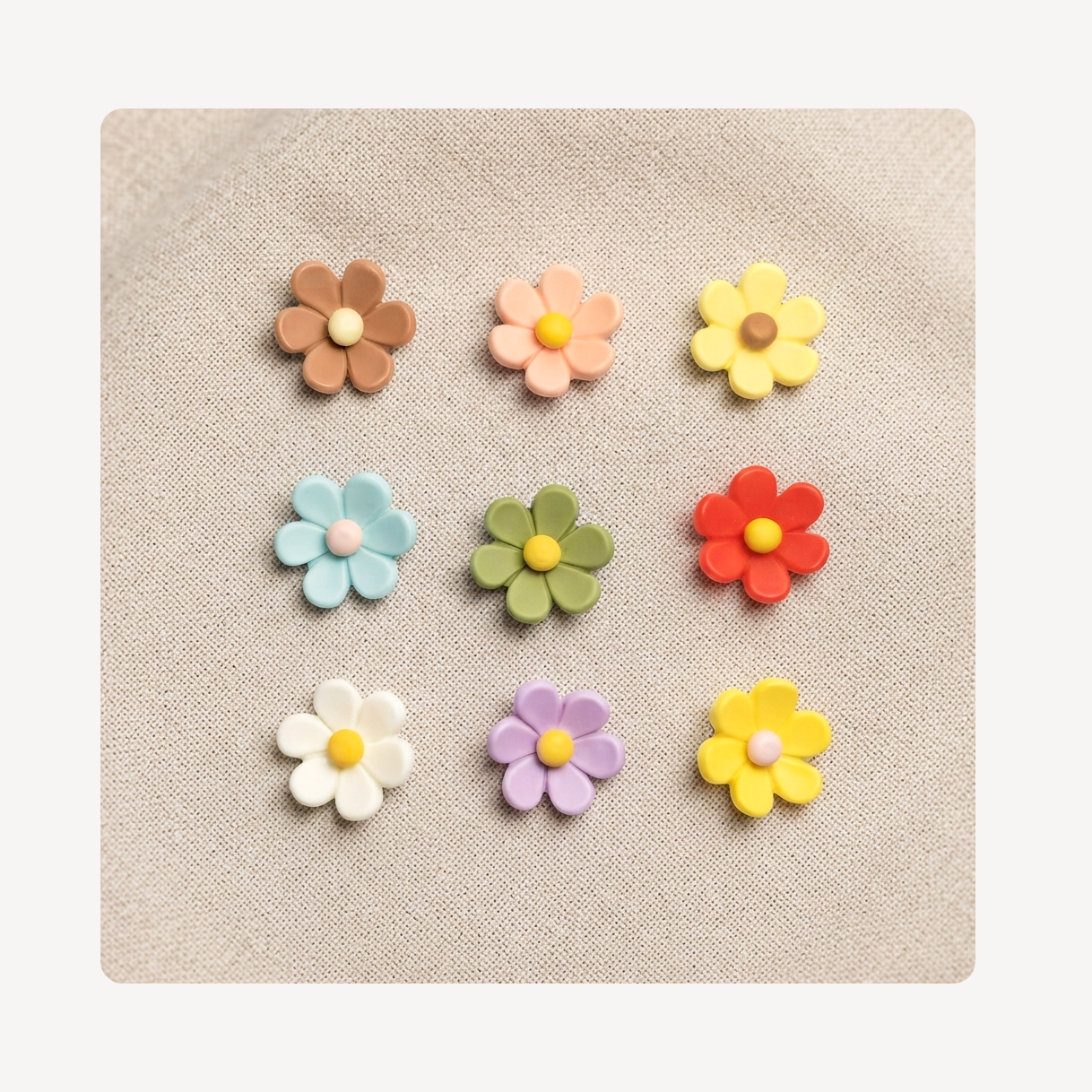 Nine small flower-shaped clog charms arranged in a 3-by-3 grid on a beige textured fabric background. The flowers come in assorted pastel and bright colors including brown, peach, yellow, light blue, green, red, white, lavender, and another yellow.