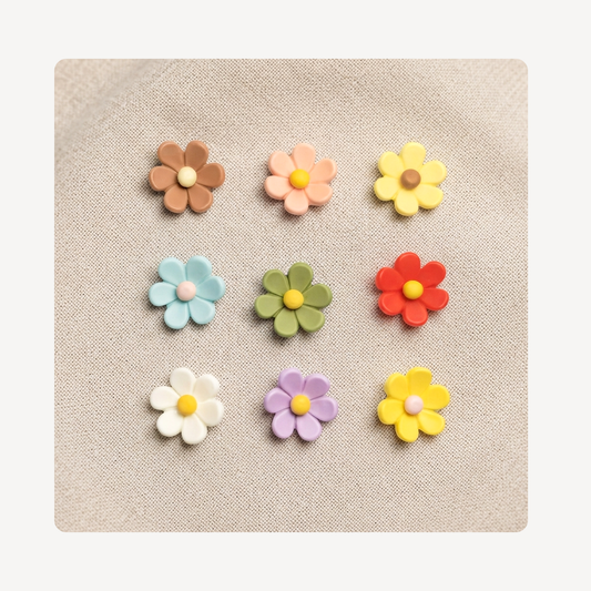 Nine small flower-shaped clog charms arranged in a 3-by-3 grid on a beige textured fabric background. The flowers come in assorted pastel and bright colors including brown, peach, yellow, light blue, green, red, white, lavender, and another yellow.