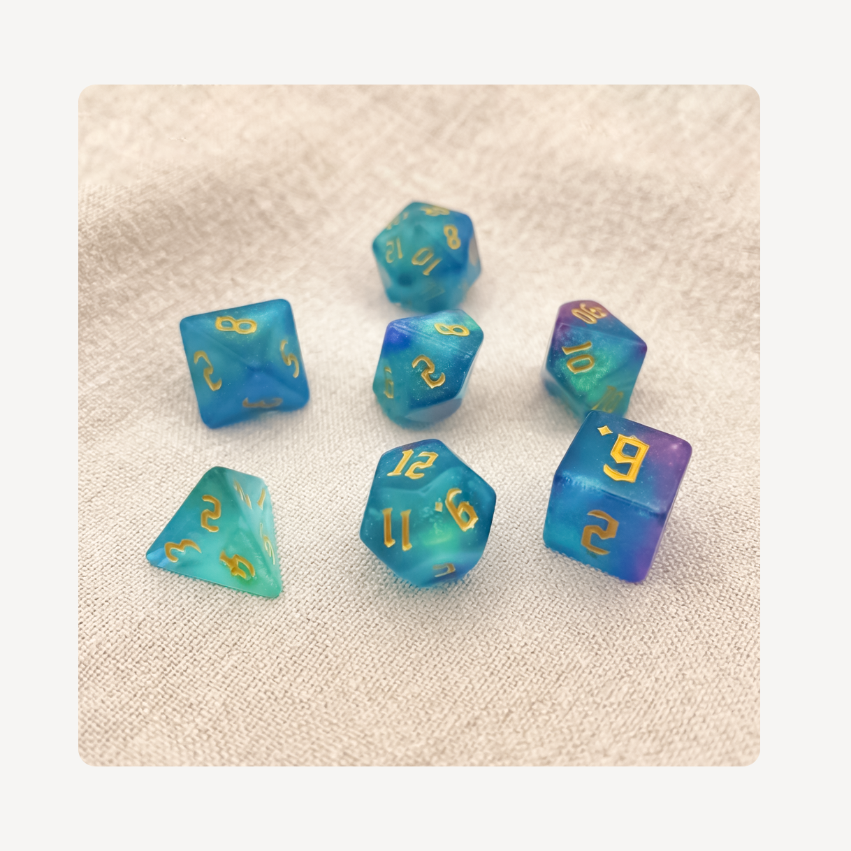 A set of seven translucent blue and teal polyhedral gaming dice clog charms  with gold numbers, arranged on a textured beige fabric background.