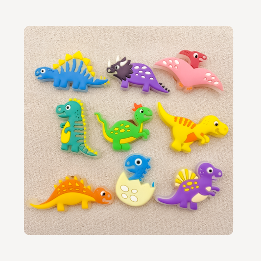 Nine colorful dinosaur-shaped clog charms arranged on a light beige textured background. The set includes various cartoon dinosaurs in blue, purple, pink, green, yellow, orange, and red, including a blue dinosaur hatching from an egg.