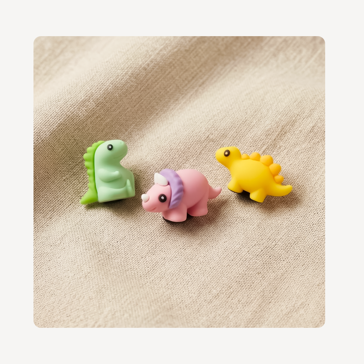Three small dinosaur-shaped clog charms in pastel green, pink, and yellow, arranged on a beige textured fabric background.