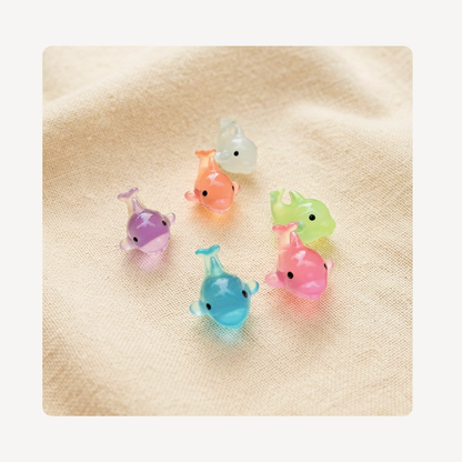 Six small translucent dolphin-shaped clog charms in pastel colors, including purple, orange, green, pink, and blue, arranged on a beige textured fabric background.