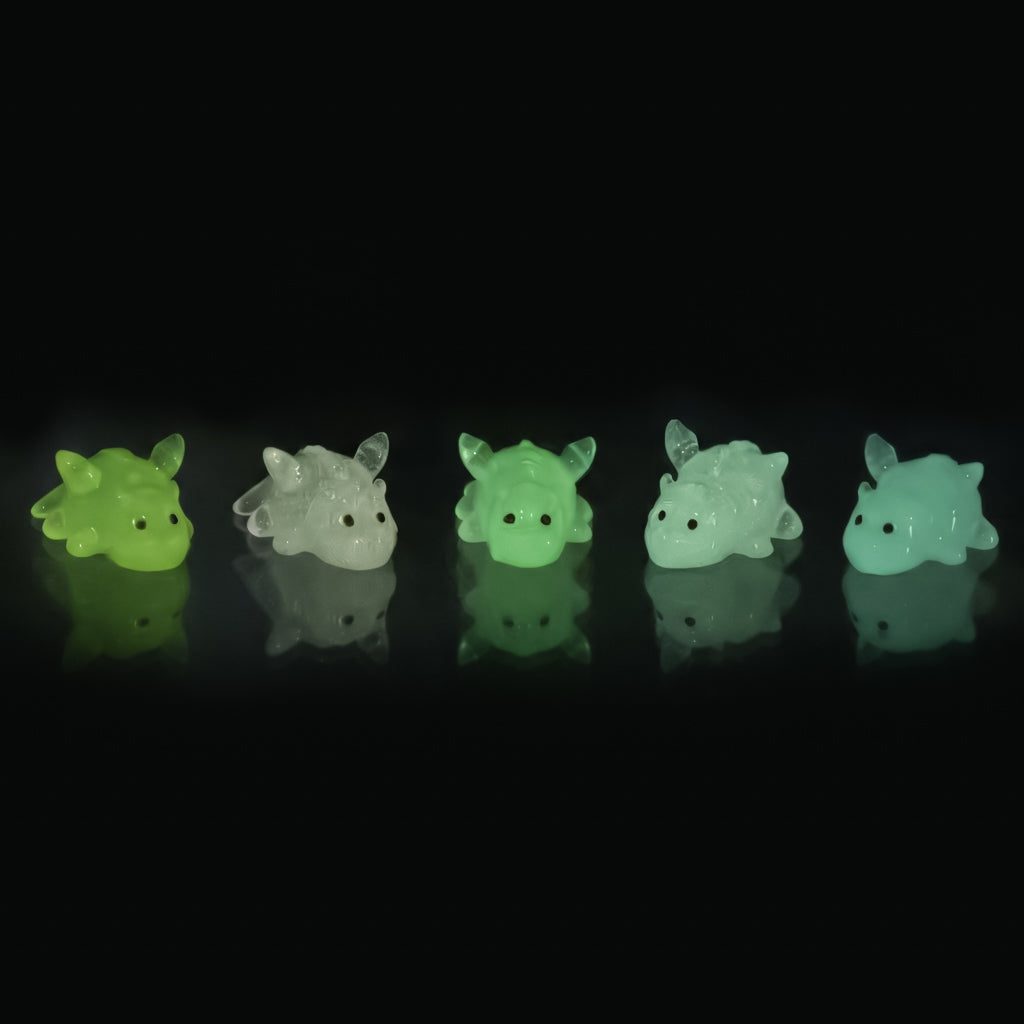 Five small dragon clog charms in assorted translucent glow-in-the-dark colors, arranged in a row on a reflective black surface against a black background.