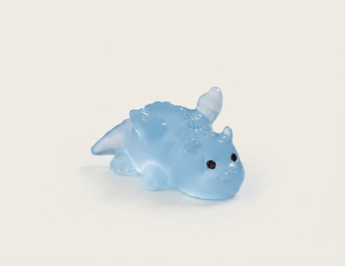 A small translucent light blue dinosaur clog charm with black eyes, photographed on a plain off-white background.