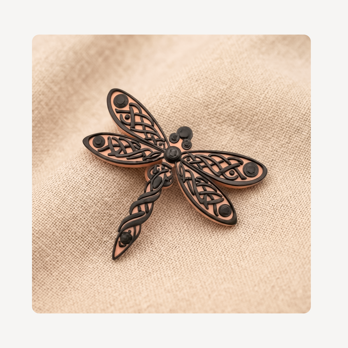 A dragonfly-shaped clog charm with intricate filigree detailing in a dark metallic finish, set against a textured beige linen background.