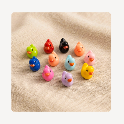 A set of small, colorful duck-shaped clog charms arranged on a textured beige fabric background. The set includes multiple colors: green, red, black, orange, pink, blue, peach, light blue, yellow, and lavender.