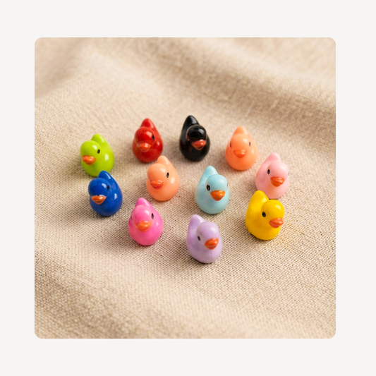 A set of small, colorful duck-shaped clog charms arranged on a textured beige fabric background. The set includes multiple colors: green, red, black, orange, pink, blue, peach, light blue, yellow, and lavender.