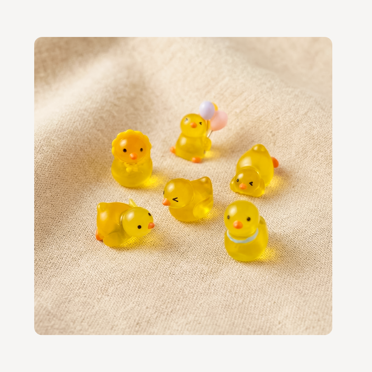 Six small translucent yellow duck clog charms with orange beaks and black eyes, arranged on a beige textured fabric background.