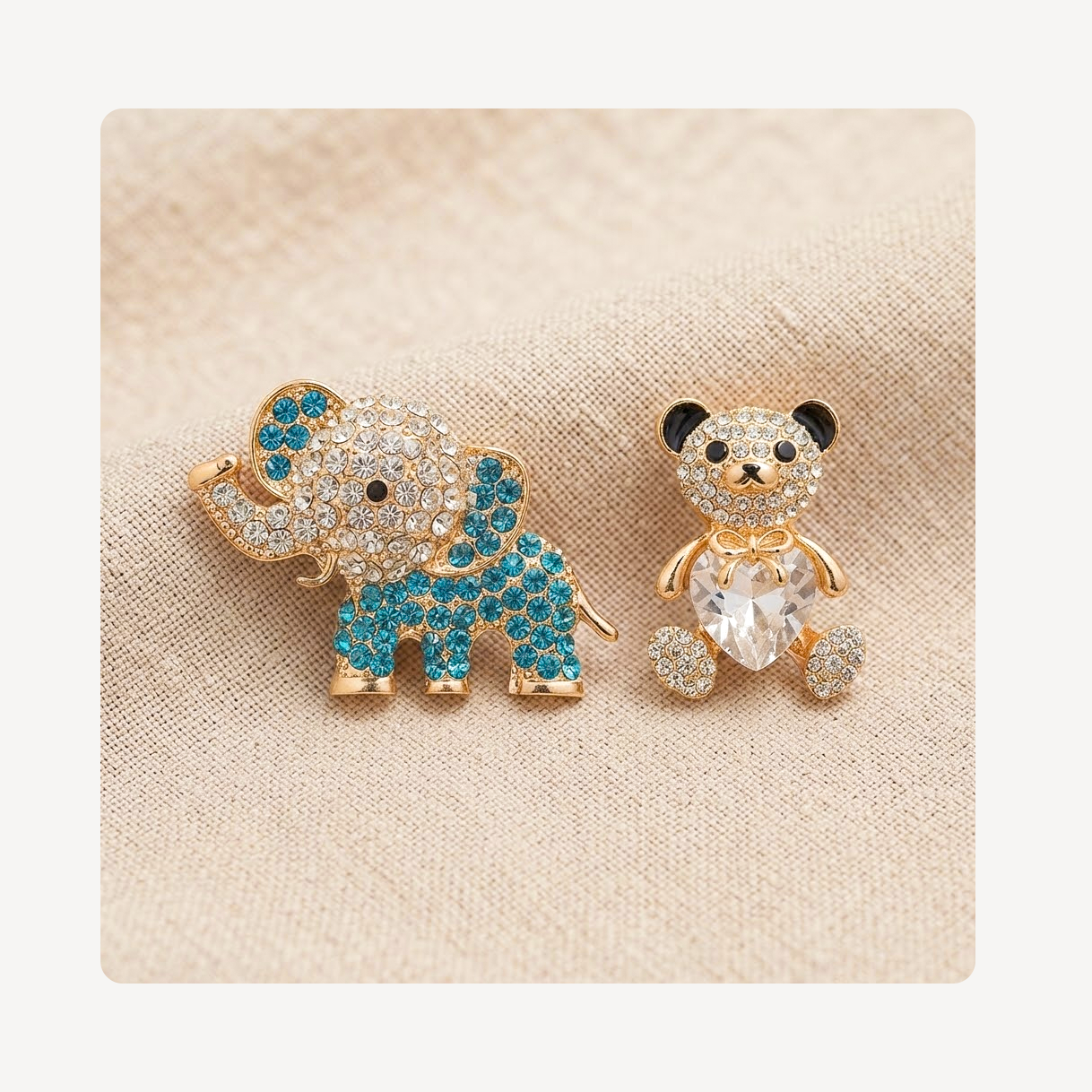 A pair of gold-tone animal clog charms, featuring a blue and clear crystal elephant on the left and a gold-tone teddy bear with a clear heart-shaped crystal on the right, displayed on a beige textured fabric background.