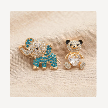 A pair of gold-tone animal clog charms, featuring a blue and clear crystal elephant on the left and a gold-tone teddy bear with a clear heart-shaped crystal on the right, displayed on a beige textured fabric background.