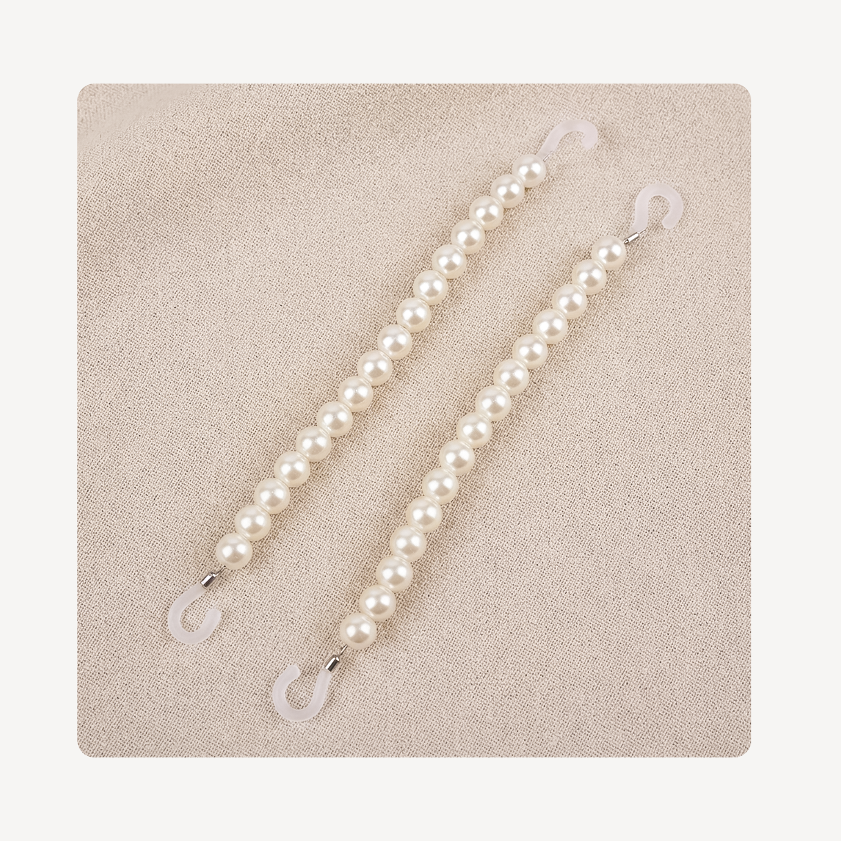 Two pearl-bead clog chains with silver-toned metal connectors and translucent white plastic hooks, displayed on a textured beige fabric background.