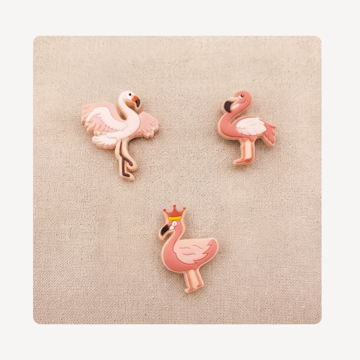 Three pink flamingo-shaped clog charms arranged on a beige textured square background, with no people or real environment visible.