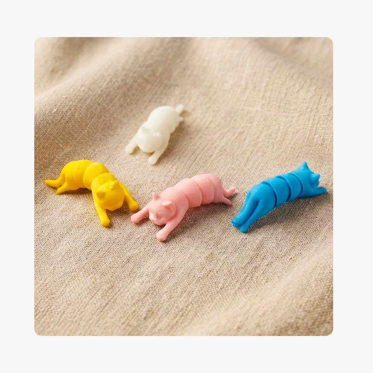 Four small cat-shaped clog charms in white, yellow, pink, and blue, arranged on a beige textured fabric background.