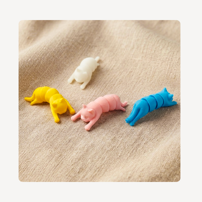 Four small cat-shaped clog charms in white, yellow, pink, and blue, arranged on a beige textured fabric background.