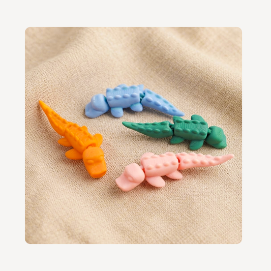 Four small crocodile-shaped clog charms in orange, light blue, green, and pink, arranged on a beige textured fabric background.