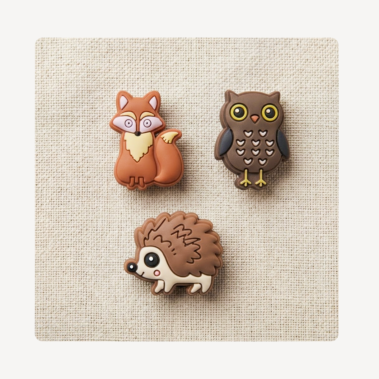 Three small animal-shaped clog charms arranged on a textured beige fabric background: a brown fox, a dark brown owl, and a hedgehog.